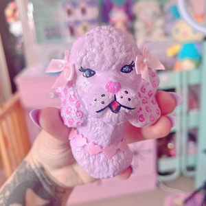 Könnte beinhalten: Eine lavendelfarbene Pudelfigur mit rosa Akzenten. Der Pudel hat ein strukturiertes Fell, einen rosa herzförmigen Kragen und kleine rosa Schleifen. Die Augen sind schwarz umrandet mit blauem Glitzer. Die Nase ist rosa und die Ohren haben rosa Flecken.