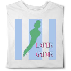 May include: A white t-shirt featuring a graphic of a green alligator silhouette centered over three vertical light blue stripes. To the right of the alligator, the text LATER GATOR is printed in pink capital letters with a black outline.