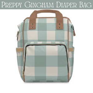 May include: A light blue and white gingham diaper bag in a backpack style. The bag has brown accents on the handle, trim, and pockets. The text "PREPPY GINGHAM DIAPER BAG" is at the top. The bag is designed for carrying baby essentials.