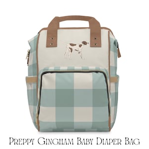 Puede incluir: Bolso de pañales para bebé con estampado de cuadros vichy en azul claro y blanco. Tiene un asa y ribetes marrones, un bolsillo delantero y un perro bordado en la parte delantera. El texto "PREPPY GINGHAM BABY DIAPER BAG" está en la parte inferior.