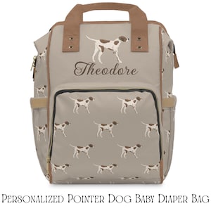 Puede incluir: Una pañalera beige con un patrón repetitivo de perros pointer marrones y blancos. La bolsa tiene asas superiores marrones, un bolsillo frontal con cremallera y el nombre Theodore impreso en letra cursiva marrón oscuro sobre el bolsillo.