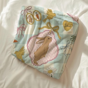 May include: A pastel blue and white checked blanket featuring illustrations of rabbits, carrots, birds, and bows. The design includes a heart with the word "Love". The blanket has a soft, pastel colour scheme.