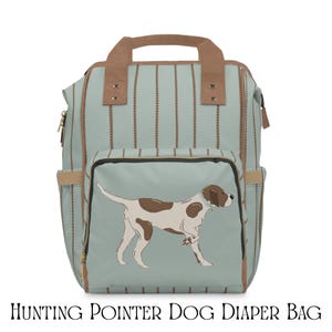 Puede incluir: Un bolso pañalera azul claro con rayas verticales marrones y detalles. El bolsillo delantero presenta una ilustración de un perro de caza. El bolso tiene un asa superior y correas ajustables. El texto "HUNTING POINTER DOG DIAPER BAG" está en la parte inferior.