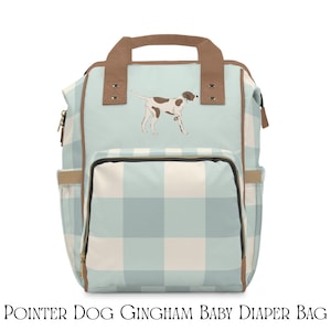 Puede incluir: Una bolsa de pañales con estampado de cuadros vichy en azul claro y blanco, con una ilustración de un perro pointer marrón y blanco en la parte delantera. Incluye asas superiores marrones, ribetes marrones, un bolsillo frontal y laterales.