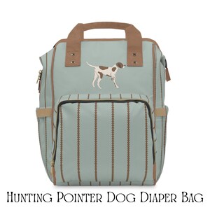 Puede incluir: Bolso de pañales azul claro con asa y detalles marrones. El bolso presenta una ilustración de perro de caza marrón y blanco y rayas verticales marrones. El texto "HUNTING POINTER DOG DIAPER BAG" está impreso en la parte inferior.