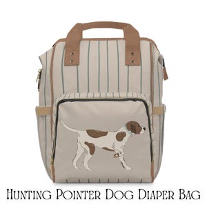 May include: A beige and brown diaper bag featuring a hunting pointer dog illustration. The bag has a striped pattern with vertical teal lines. It includes a top handle, side pockets, and a front pocket with the dog graphic. The text "HUNTING POINTER DOG DIAPER BAG" is at the bottom.