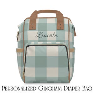 Puede incluir: Un bolso de pañales de cuadros vichy azul claro y blanco con detalles en marrón. El bolso tiene un asa superior, múltiples bolsillos y el nombre "Lincoln" en cursiva. El texto "PERSONALIZED GINGHAM DIAPER BAG" está en la parte inferior.