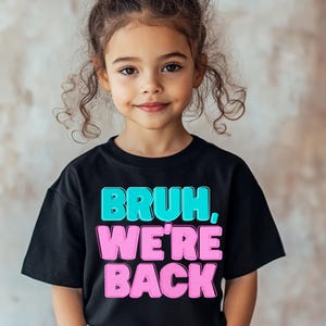 May include: A black t-shirt with the text "BRUH, WE'RE BACK" in turquoise and pink bubble letters. The shirt is worn by a young girl with curly brown hair.
