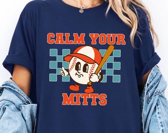 May include: A navy blue short-sleeve t-shirt featuring a graphic of a cartoon baseball wearing a cap and holding a bat. The text CALM YOUR MITTS is printed in orange above and below the baseball, set against a teal and navy checkered background.