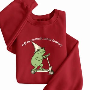 May include: A red sweatshirt featuring a cartoon frog on a scooter, wearing a white hat, with the text "Off to commit mom foolery". The frog is green. The sweatshirt is a solid, deep red colour.