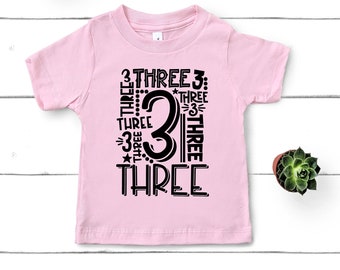 May include: Pink short-sleeved t-shirt with black text that says "3 THREE 3: THREE 3 THREE THREE 3 THREE THREE" with a large black number 3 in the center.