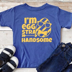 May include: A blue t-shirt with a yellow graphic of an egg wearing sunglasses and a bow tie. The text "I'm egg-stra handsome" is printed on the shirt.