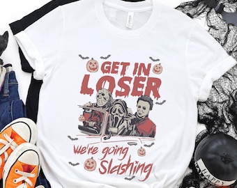 May include: White t-shirt with a Halloween graphic featuring horror movie characters and the text "Get in Loser, we're going slashing." The shirt is paired with orange sneakers, jeans, and Halloween decorations.