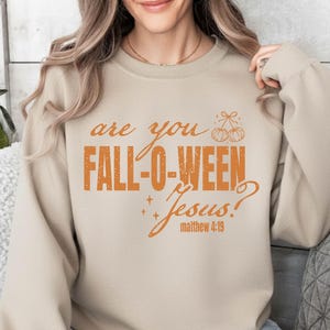 May include: Beige sweatshirt with the text "are you FALL-O-WEEN Jesus?" in orange. The text includes a pumpkin graphic and the Bible verse Matthew 4:19. The sweatshirt is a neutral color and suitable for fall and Halloween.