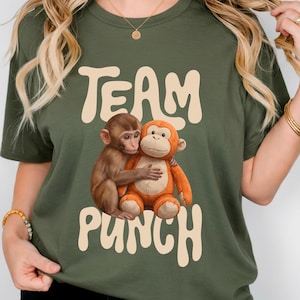 May include: Olive green t-shirt with the words "TEAM PUNCH" in a retro font. The shirt features a graphic of two monkeys hugging. One monkey is brown, and the other is orange.