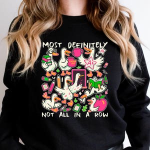 May include: Black sweatshirt with a colorful graphic of cartoon ducks with books, pencils, and apples. The text on the sweatshirt reads "MOST DEFINITELY NOT ALL IN A ROW."