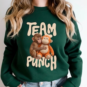 May include: Dark green sweatshirt with the text "TEAM PUNCH" in a cream-colored, retro font. The sweatshirt features a graphic of two hugging monkeys, one brown and one orange.