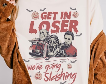 May include: Cream-colored long-sleeve shirt with a Halloween-themed graphic. The graphic features the text "GET IN LOSER" above a car with Jason Voorhees, Ghostface, and Michael Myers. Below the car is the text "we're going slashing".