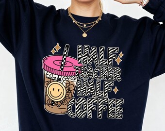 May include: A navy blue crewneck sweatshirt featuring a graphic print of an iced coffee cup with a pink lid, a straw, and a yellow smiley face icon. The text reads "HALF TEACHER HALF COFFEE" in a black and white zebra-striped font with star accents.