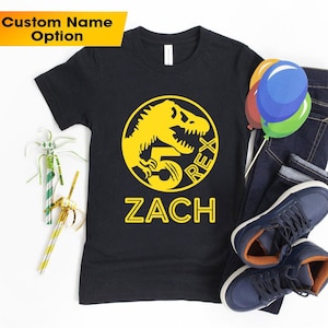 Op de afbeelding: Zwart T-shirt met een gele dinosaurus afbeelding en de tekst "5 REX ZACH" erop.