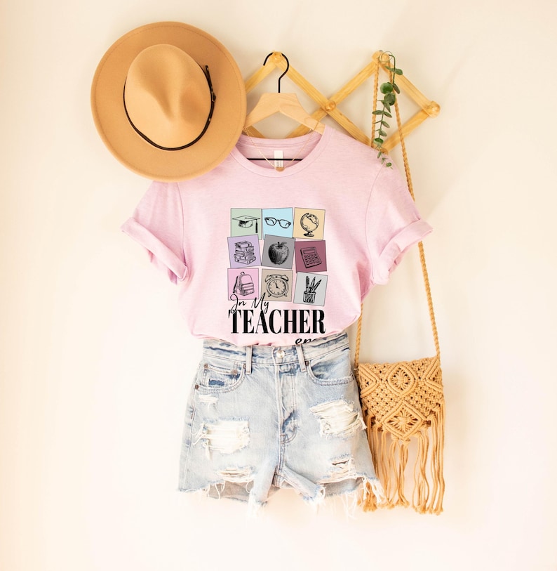 May include: Pink t-shirt with a graphic of school supplies in colorful squares and the text "In My Teacher Era".