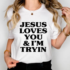 May include: White t-shirt with the text "JESUS LOVES YOU & I'M TRYIN" in bold black letters. The shirt is short-sleeved and appears to be made of a soft material. The person is wearing gold bracelets and a necklace.