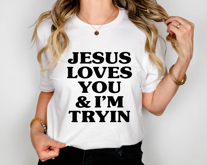 May include: White t-shirt with the text "JESUS LOVES YOU & I'M TRYIN" in bold black letters. The shirt is short-sleeved and appears to be made of a soft material. The person is wearing gold bracelets and a necklace.