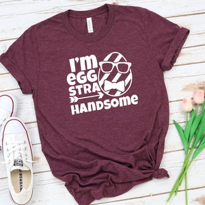 May include: A maroon t-shirt with a white graphic of an egg wearing sunglasses and a bow tie. The text on the shirt reads "I'm egg-stra handsome".