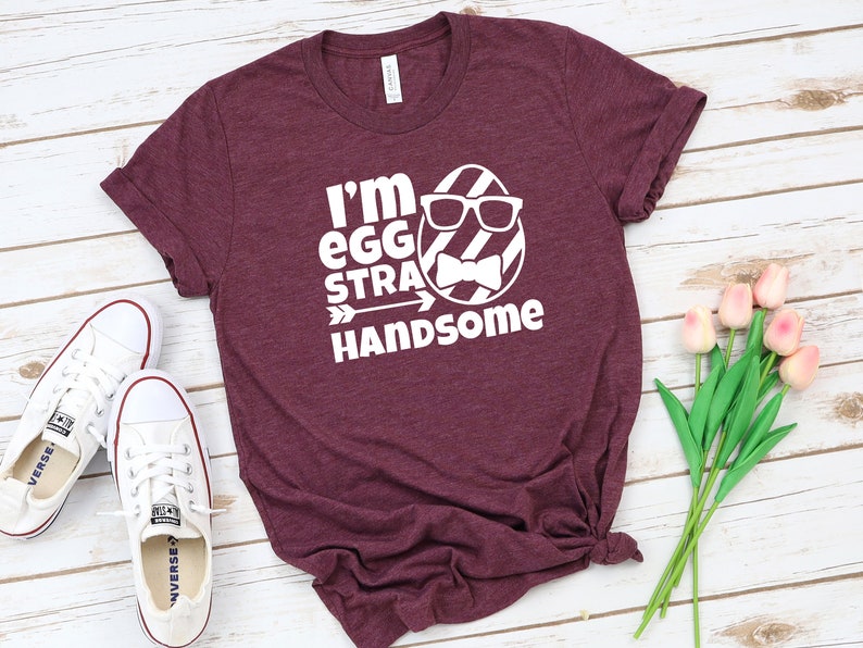 May include: A maroon t-shirt with a white graphic of an egg wearing sunglasses and a bow tie. The text on the shirt reads "I'm egg-stra handsome".