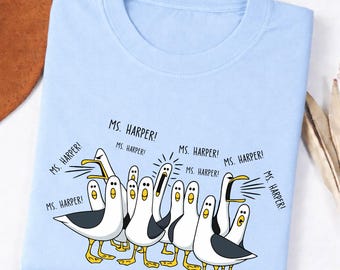 May include: A light blue t-shirt featuring a graphic of multiple white and gray seagulls with yellow beaks and feet. The seagulls are depicted with open beaks, and the text "MS. HARPER!" is printed repeatedly in black above and around the birds.