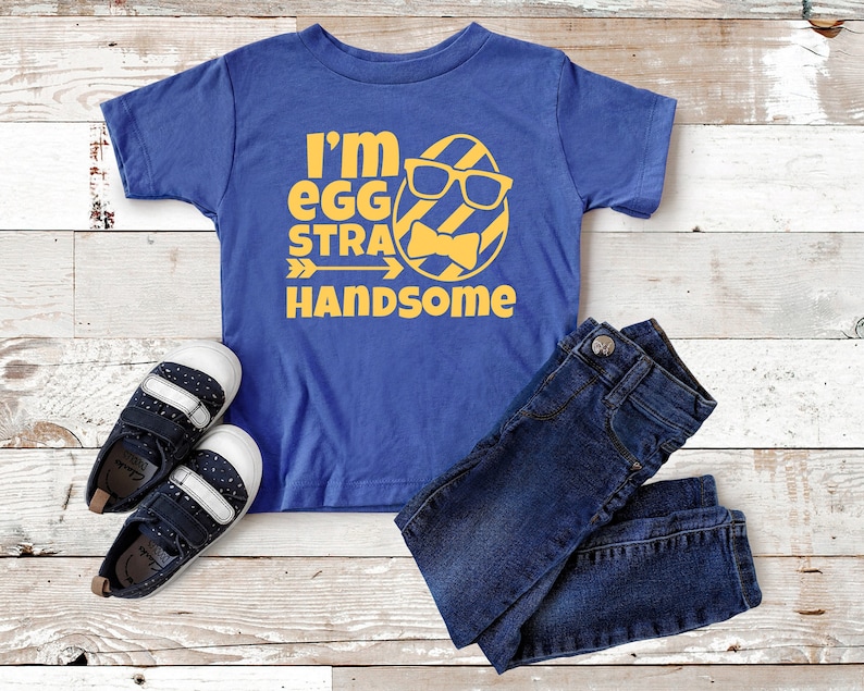 May include: A blue t-shirt with a yellow graphic of an egg wearing sunglasses and a bow tie. The text "I'm egg-stra handsome" is printed on the shirt.