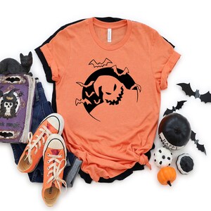 May include: Orange t-shirt featuring a black silhouette of a ghost with bats flying around it. The ghost is in a crescent moon shape.