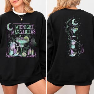 May include: A black long-sleeve sweatshirt featuring a graphic print with the text MIDNIGHT MARGARITAS. The design includes a margarita glass, a moka pot, a frog wearing a purple hat, candles, and a crescent moon in shades of green and purple.