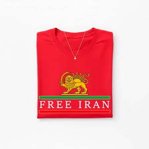 Może przedstawiać: Złożona czerwona koszulka z naszyjnikiem ze złotą gwiazdą. Koszulka ma złoty emblemat lwa i słońca z napisem "FREE IRAN" białymi literami na czerwonym tle z zielonymi i białymi paskami.