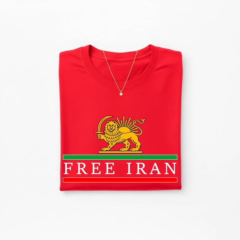 Może przedstawiać: Złożona czerwona koszulka z naszyjnikiem ze złotą gwiazdą. Koszulka ma złoty emblemat lwa i słońca z napisem "FREE IRAN" białymi literami na czerwonym tle z zielonymi i białymi paskami.