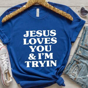 May include: Royal blue t-shirt with the text "JESUS LOVES YOU & I'M TRYIN" in white. The shirt is displayed with white sneakers, denim shorts, and heart-shaped sunglasses on a wooden surface.