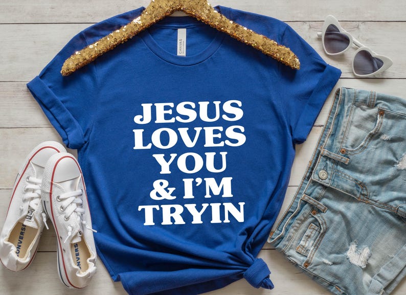 May include: Royal blue t-shirt with the text "JESUS LOVES YOU & I'M TRYIN" in white. The shirt is displayed with white sneakers, denim shorts, and heart-shaped sunglasses on a wooden surface.