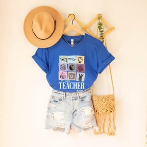 May include: A royal blue t-shirt with a graphic of a grid of school supplies, including a graduation cap, glasses, a globe, books, an apple, a calculator, a backpack, a clock, a pencil, and a ruler. The text "In My Teacher Era" is printed below the graphic.
