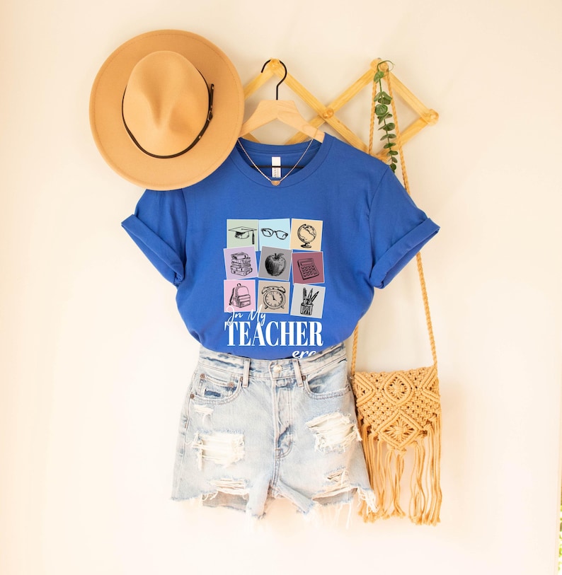 May include: A royal blue t-shirt with a graphic of a grid of school supplies, including a graduation cap, glasses, a globe, books, an apple, a calculator, a backpack, a clock, a pencil, and a ruler. The text "In My Teacher Era" is printed below the graphic.