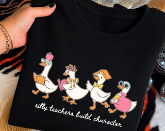 May include: A black crewneck sweatshirt featuring a graphic of four white ducks dressed as teachers. The ducks wear accessories like glasses, a flower crown, and a book on their heads. Below the ducks is the white text, silly teachers build character.