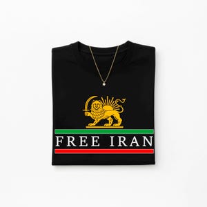 Może przedstawiać: Czarny t-shirt ze złotym lwem i słońcem, zielono-czerwonym paskiem i napisem "FREE IRAN". Złoty naszyjnik z wisiorkiem w kształcie gwiazdy jest zarzucony na koszulkę.