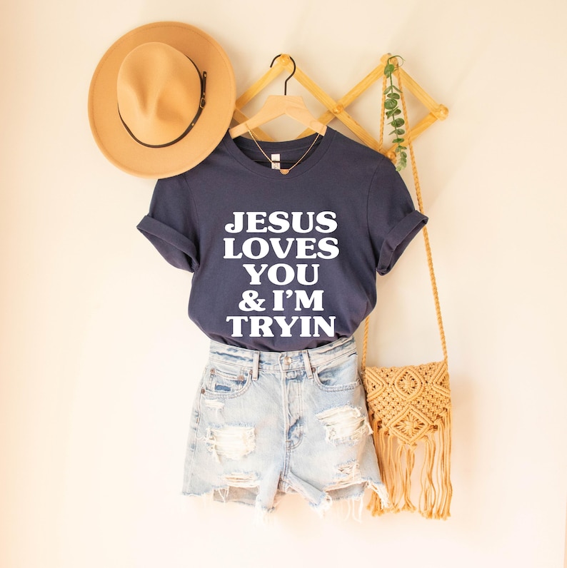 May include: Navy blue t-shirt with the text "JESUS LOVES YOU & I'M TRYIN" in white. The shirt is paired with light wash denim shorts. A tan hat and a woven bag with fringe are also visible.