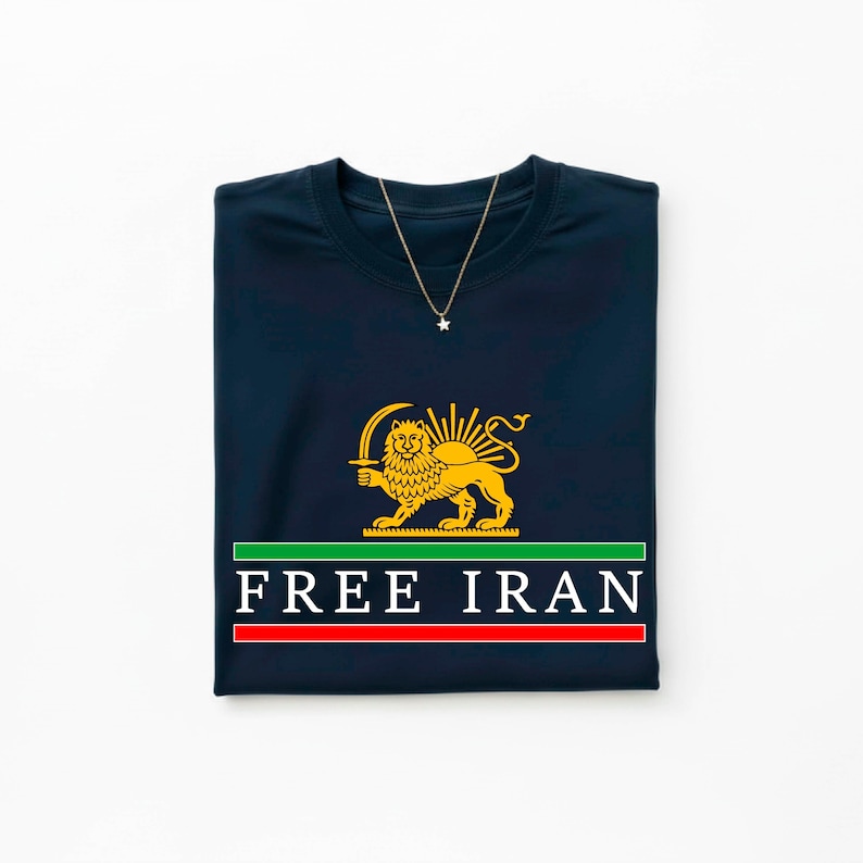 Może przedstawiać: Granatowa koszulka z grafiką złotego lwa i miecza, zielono-czerwonym paskiem i napisem "FREE IRAN". Złoty naszyjnik z wisiorkiem w kształcie gwiazdy jest zarzucony na koszulkę.