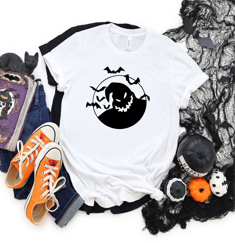 May include: White t-shirt featuring a black silhouette of a grinning ghost with bats flying around it. The ghost is in a circle, resembling a full moon.