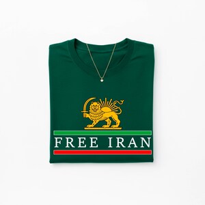 Może przedstawiać: Złożona ciemnozielona koszulka z złotą grafiką lwa trzymającego miecz i białym napisem "FREE IRAN". Złoty naszyjnik z wisiorkiem w kształcie gwiazdy jest zarzucony na koszulkę.
