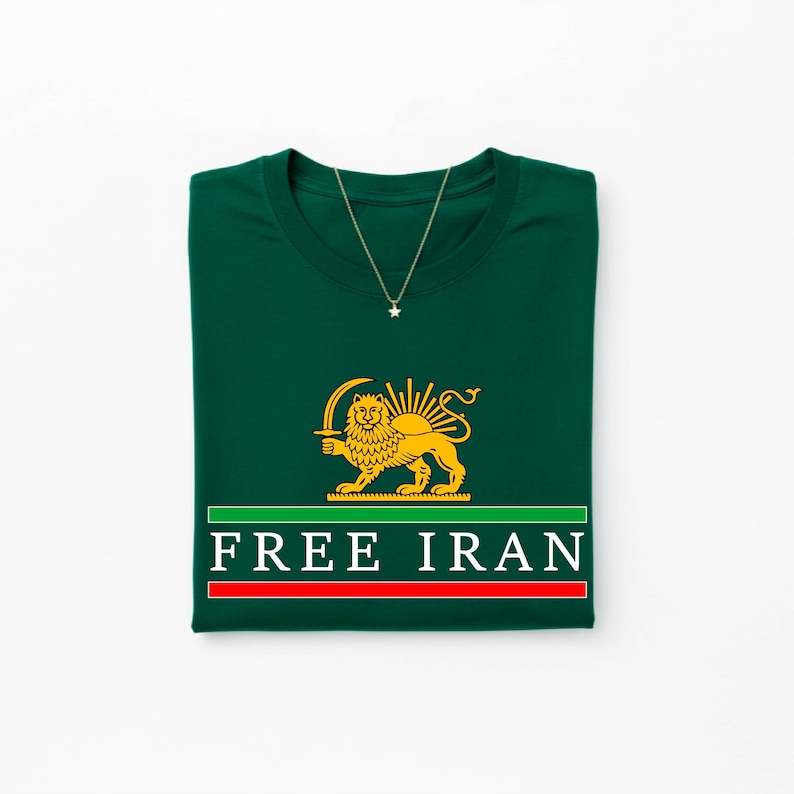 Może przedstawiać: Złożona ciemnozielona koszulka z złotą grafiką lwa trzymającego miecz i białym napisem "FREE IRAN". Złoty naszyjnik z wisiorkiem w kształcie gwiazdy jest zarzucony na koszulkę.