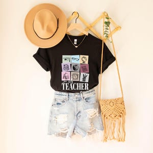 May include: Black t-shirt with a graphic of colorful squares featuring school supplies and the text "For My Teacher". The shirt is displayed on a hanger with a straw hat and a woven bag.