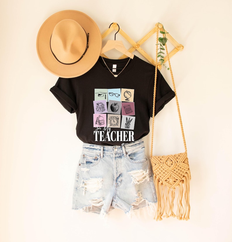 May include: Black t-shirt with a graphic of colorful squares featuring school supplies and the text "For My Teacher". The shirt is displayed on a hanger with a straw hat and a woven bag.