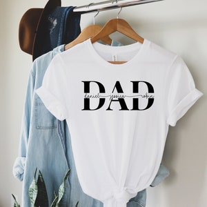 May include: White t-shirt with the word "DAD" in bold black letters. The names "Daniel", "Jessica", and "Robin" are written in cursive script above and below the letters.