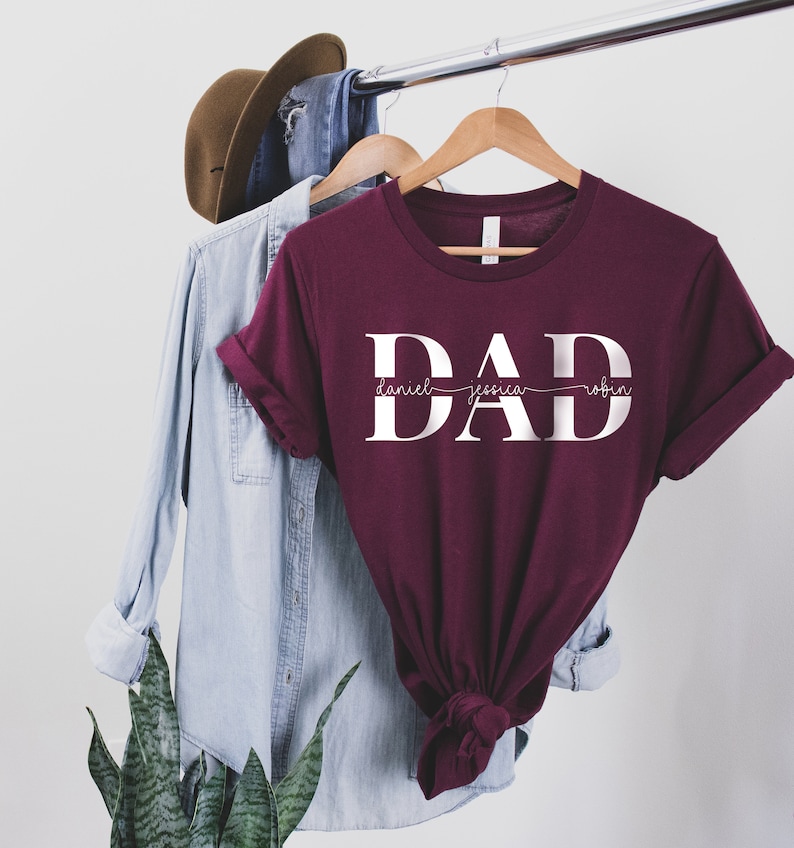 May include: Burgundy t-shirt with the word "DAD" in white letters. The names "Daniel", "Jessica", and "Robin" are written in a smaller font underneath the word "DAD".